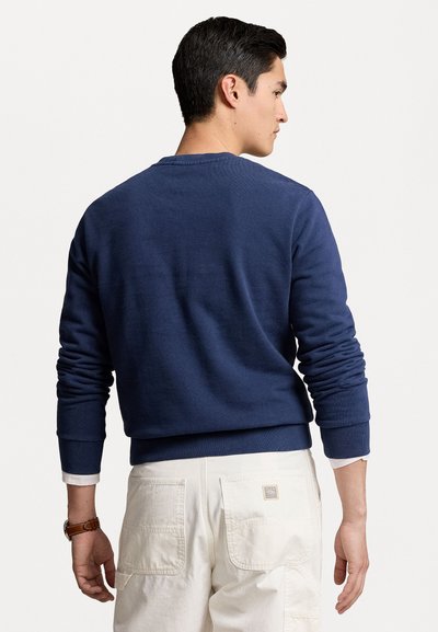Tumedat sinise värviga crewneck sviiter ribitud varrukate ja allääriga, valmistatud pehmest kangast. Mudel kannab heledat värvi pükse ja kella.