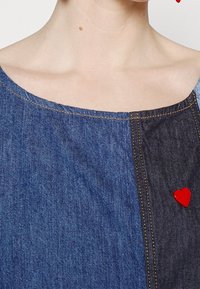 Denimtopp med båtformad halsringning, kontrasterande blå och mörka denimpaneler samt en röd hjärtformad nål på vänster sida.