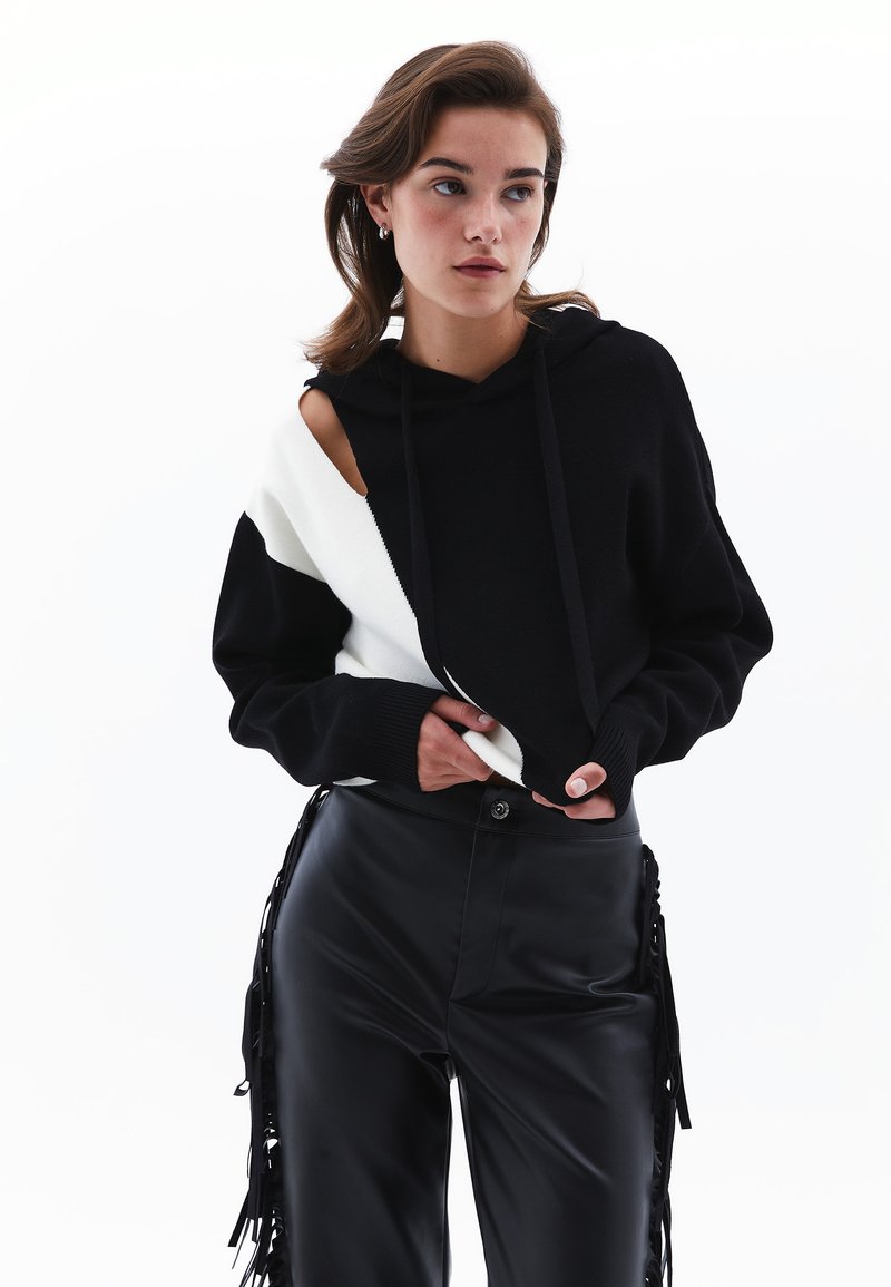 Schwarzer und weißer Kapuzenpullover mit ausgeschnittenem Schulterdesign und Zugbändern, kombiniert mit schwarzen Kunstlederhosen mit Fransenakzenten.
