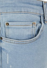 Poche de jean en denim bleu clair avec rivets en cuivre visibles et coutures orange, montrant la texture du tissu et des marques d'usure légères.