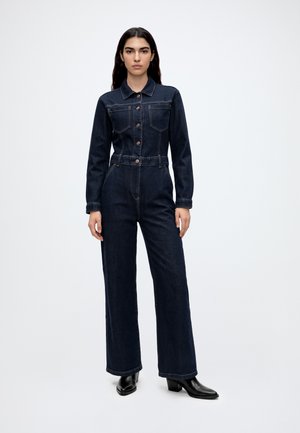 Une femme porte une combinaison en jean foncé, avec un haut boutonné de style veste et des jambes larges, associée à des chaussures noires à talons, sur un fond uni.