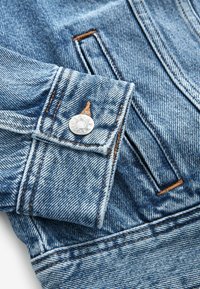 Detalle de una chaqueta de denim azul claro que presenta un cierre de botones, costuras en contraste y un puño doblado con un bolsillo visible. Tejido texturizado.