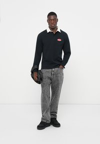 Black long-sleeve polo shirt with a white collar, red logo patch. Worn with grey patterned loose-fit jeans and black shoes, holding a jacket.