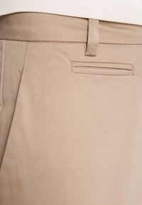 Beiga byxor med en slät textur, featuring en bakficka och en flat frontdesign, med sömdekoration längs sömmarna.