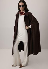 Sieviete, kas valkā tumši brūnu apmetni virs baltas tunikas un platajiem biksēm, ar brūniem garajiem apaviem, sarkanu rakstainu šalli, saulesbrillēm un zelta aproci.