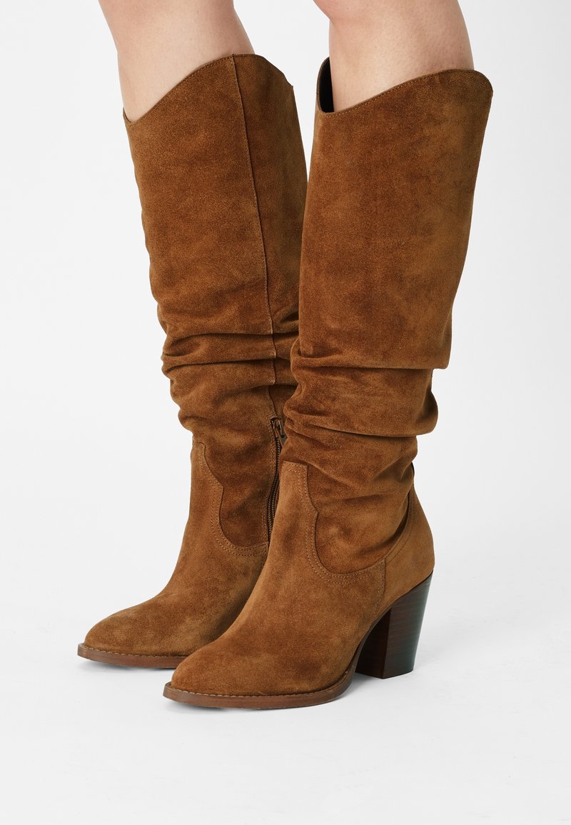 Brązowe, zamszowe kozaki za kolano z luźnym fasonem, szpiczastym noskiem i piętą na słupku. Wyposażone w zamek błyskawiczny z boku dla łatwego zakładania.