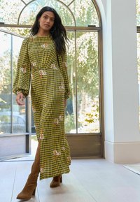 Olīvu zaļš maxi kleita ar rūtainu rakstu, ziedu akcentiem, garām pūkainām piedurknēm un sānu šķēlumu, apvienota ar brūnām zamšādām potītes zābakiem.