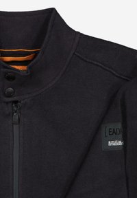 Black fleece jacket with a high collar, zip closure, and two button accents. Features an orange inner lining and a label on the side.