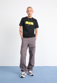 Black T-shirt with "AIR" graphic in yellow, paired with light gray cargo pants featuring side pockets and white sneakers with black accents.