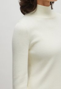 Weißer Rollkragenpullover aus weichem Strickstoff, mit tailliertem Schnitt und geripptem Muster entlang des Kragens und der Manschetten.
