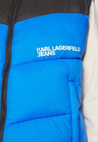 Blå och svart vadderad jacka med frontdragkedja och vitt "KARL LAGERFELD JEANS"-logotyp på bröstet, bärs över en krämfärgad långärmad tröja.