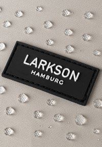Textúrált bézs szövet, kiemelkedő ezüst pöttyökkel, amelyen alul egy fekete téglalap alakú címke található, fehér "LARKSON HAMBURG" felirattal.