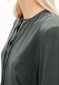 Tummanvihreä pusero, jossa on pehmeä, silkkimäinen koostumus, pyöreä pääntie avattavalla riipuksella ja yksittäinen kaulanauhan yksityiskohta.