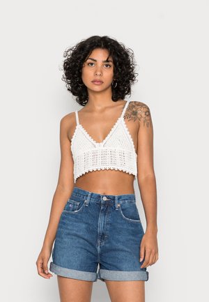 Top cropped de croché branco com bordas em forma de onda, combinado com calções de denim azul de cintura alta e bainha enrolada. Textura da pele visível, design simples.