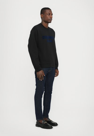 Sweat-shirt noir avec logo bleu, jeans en denim et mocassins noirs. Le mannequin se tient de côté devant un fond neutre.