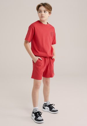 Completo in cotone rosso composto da t-shirt e pantaloni corti, con scollatura rotonda e tasche laterali. Presenta un logo discreto sulla maglietta. Abbinato a sneakers nere e bianche.