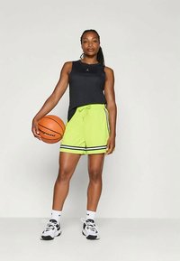 Schwarzes ärmelloses Oberteil, neongrüne Shorts mit schwarzen Streifen, Basketball in der Hand, weiße und schwarze Sneaker. Neutraler Hintergrund.