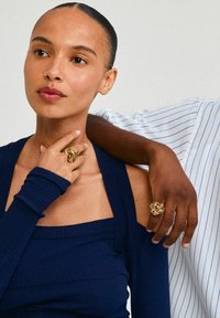 Haut à manches longues côtelé marine avec un décolleté carré, présentant une bague texturée dorée sur une main et une main sombre portant une bague avec une fleur.
