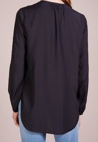 Blouse noire à manches longues avec un col rond, un détail froncé à l'arrière et une coupe décontractée, associée à un jean bleu clair.