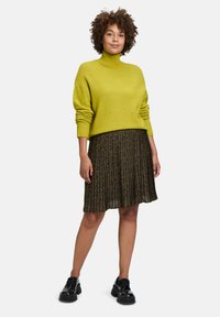 Lindenfarbener gestrickter Rollkragenpullover, kombiniert mit einem schwarzen gemusterten gefalteten Rock und schwarzen Schnürschuhen.