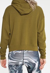 Kvinna med blont hår iklädd en olivgrön hoodie och leggings med svart-vitt abstrakt mönster, ses bakifrån.