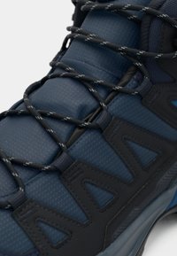 Marineblå sportssko med svarte mønstrede snører som går gjennom svarte trekantede øyelet, med teksturert stoff og polstret tunge.