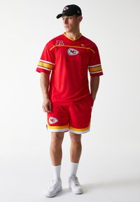 Piros hálós sportme sárga és fehér csíkokkal, csapattal logóval. Hozzá illő piros rövidnadrág és fekete sapka. Fehér sportcipő.