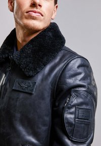 Veste bomber en cuir noir avec un col en fourrure noire douce, surface texturée, poches latérales zippées et un insigne sur la poitrine.