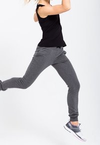 Osoba w powietrzu nosi czarny sportowy tank top i szare spodnie dresowe, do tego szaro-różowe buty do biegania, na białym tle.