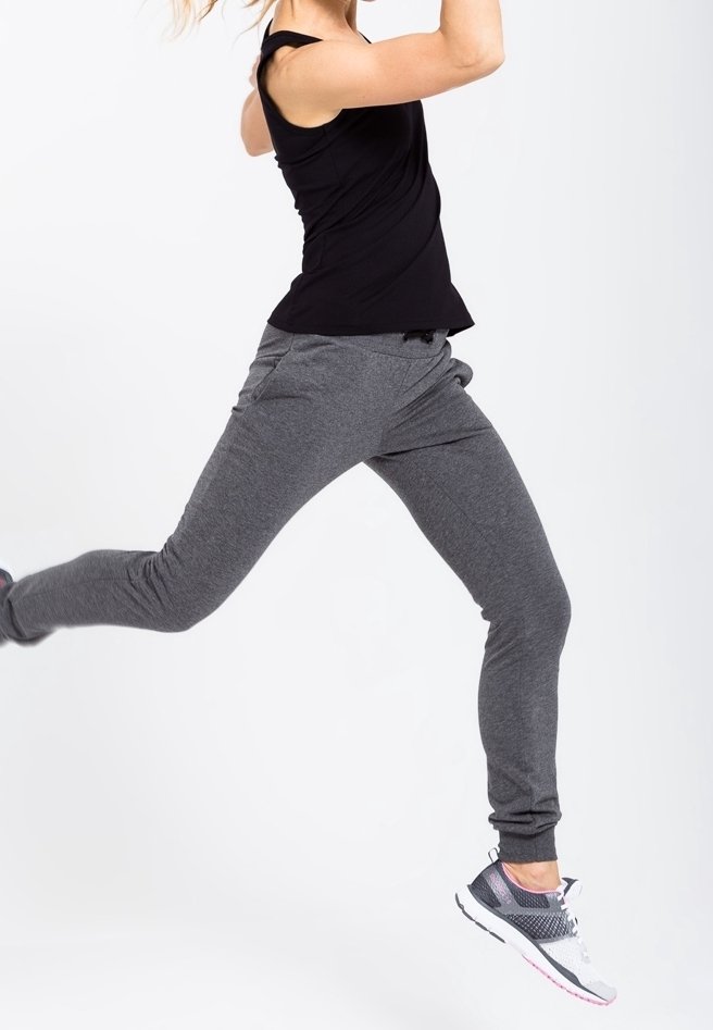 Osoba w powietrzu nosi czarny sportowy tank top i szare spodnie dresowe, do tego szaro-różowe buty do biegania, na białym tle.