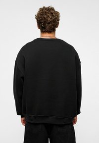 Zwarte sweatshirt met een ontspannen pasvorm, verlaagde schouders en brede manchetten. De stof is zacht, met een gladde textuur en subtiele stiksels.