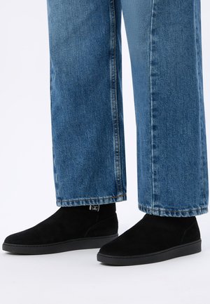 Person wearing black suede ankle boots and straight-leg blue denim jeans against a white background.