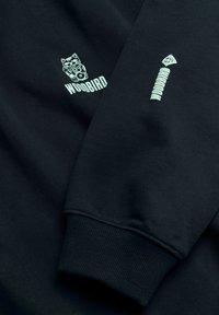 Manches de sweat-shirt noires avec un logo de chat au-dessus du mot "WOODBIRD" et un patch vertical de formes empilées stylisées sur l'autre manche.