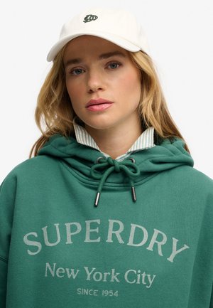 Jonge vrouw draagt een witte pet en een tealkleurige Superdry hoodie met aantrekkoorden over een gestreept overhemd met kraag, kijkt recht vooruit.
