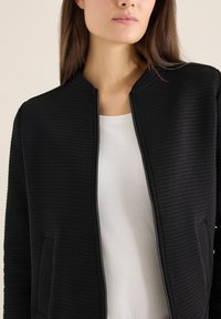 Giacca nera con trama, con zip frontale, collo rotondo e tasche laterali; indossata sopra una camicia bianca.