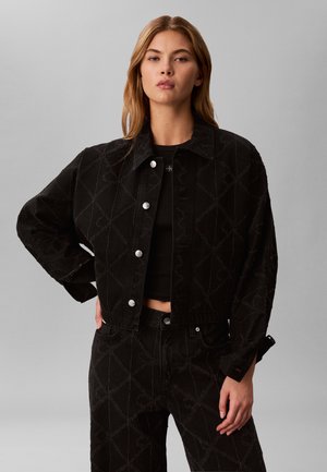 Ung kvinde med blondt hår modellerer sort mønstret jakke og matchende bukser med sølvknapper mod ensartet gråt baggrund.