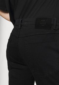 Svart denimjeans som bärs med en svart skjorta, med fokus på bakfickan och lädermärket med "NEUW Stockholm Melbourne 2009."