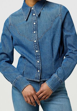 Kobieta ubrana w dopasowaną niebieską kurtkę dżinsową na zatrzaski oraz pasujące niebieskie dżinsy, ręce luźno splecione na talii.