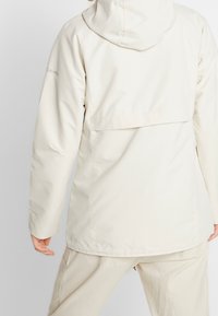 Osoba v krémově bílé voděodolné bundě s kapucí a odpovídajících kalhotách, stojící zády k fotoaparátu na jednobarevném pozadí.