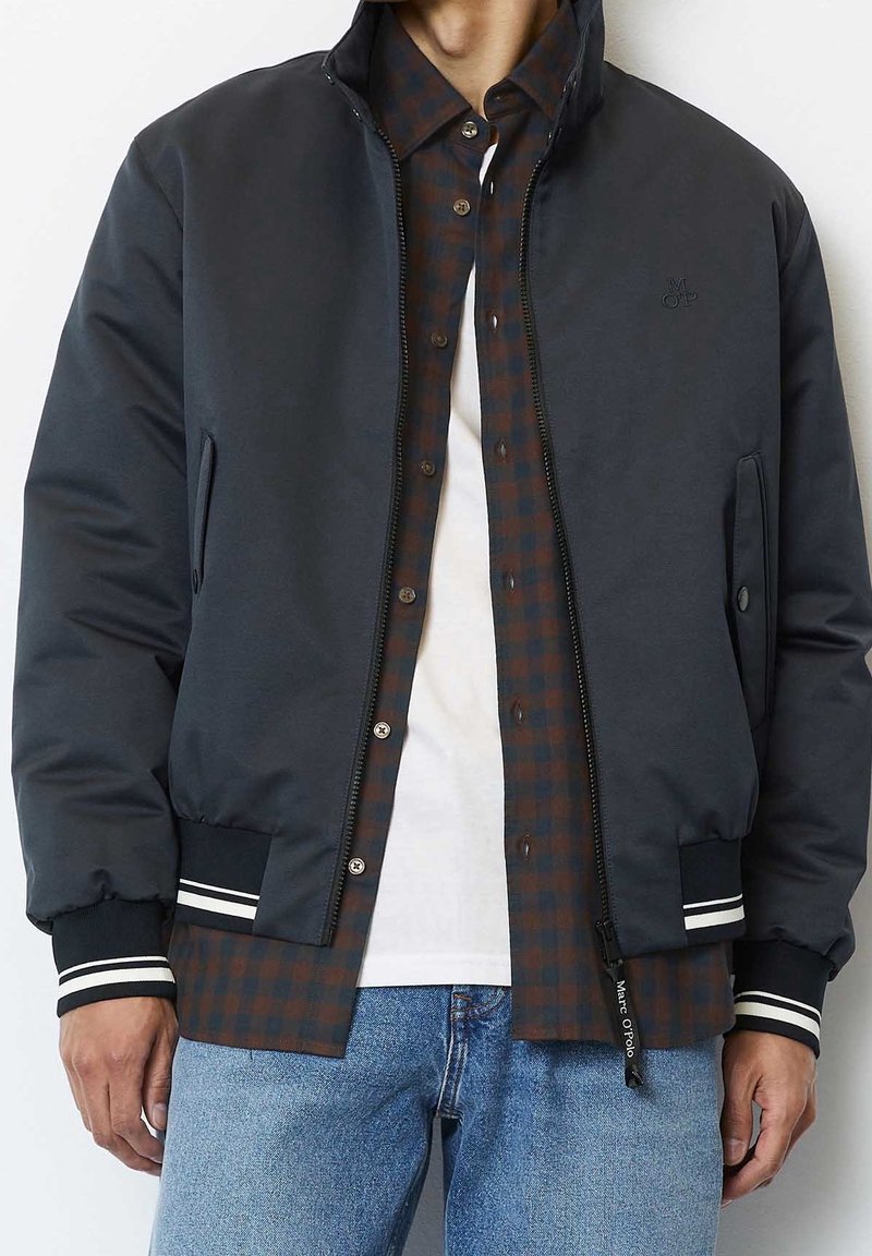 Giacca bomber blu navy scuro con collo a coste, dettagli a strisce bianche e tasche laterali, indossata sopra una camicia marrone a quadri e una t-shirt bianca.