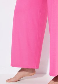 Růžové široké kalhoty vyrobené z měkké látky. Mají volný střih, hladkou texturu a prošité lemy. Skvěle ladí s bosou nohou v kontextu stylingu.
