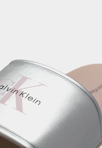 Ασημί σαγιονάρα Calvin Klein με μεταλλική υφή και ανοιχτό ροζ σόλα, εμφανίζοντας μερικό λογότυπο της μάρκας στο λουράκι.