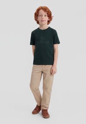 Garçon roux debout, portant un t-shirt vert foncé "ORIGINALS", un pantalon beige et des baskets marron sur un fond clair uni.