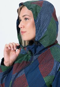 Žena upravující kapuci barevné bundy s puntíkovaným vzorem v modré, zelené, červené a bílé barvě na jednobarevném pozadí.