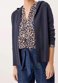 Nainen, joka käyttää tummansinistä hupparia monivärisen kuviollisen paidan päällä, joka on piilotettu korkeavyötäröisiin sinisiin farkkuihin, joissa on solmittu vyö.