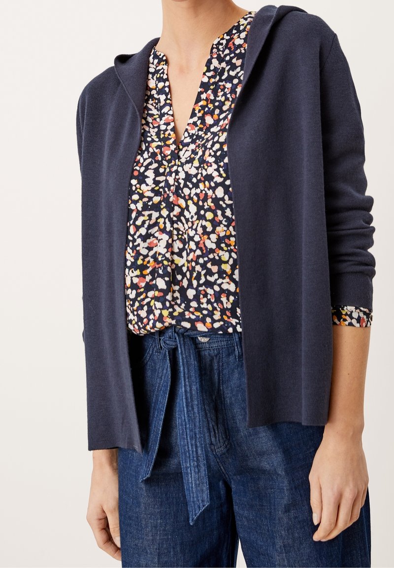 Nainen, joka käyttää tummansinistä hupparia monivärisen kuviollisen paidan päällä, joka on piilotettu korkeavyötäröisiin sinisiin farkkuihin, joissa on solmittu vyö.