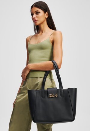 Czarna skórzana torba tote w zelektryzowanym kształcie, z złotym logo i długimi uchwytami. Modelka ma na sobie zielony top o fakturowanym wzorze.