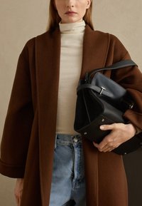 Cappotto di lana marrone con maniche ampie, dolcevita color crema, jeans a vita alta azzurri chiari e una borsa in pelle nera con accessori in argento.