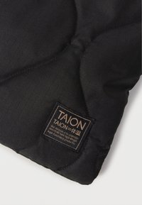 Crni quilted materijal s suptilnim kariranim uzorkom, s pravokutnom oznakom na kojoj je prikazano ime brenda "TAION" i misijska izjava.