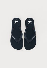 Tongs bleu marine avec semelles texturées et fines lanières. Présente le texte et le logo "Calvin Klein Jeans" sur la semelle et la lanière.